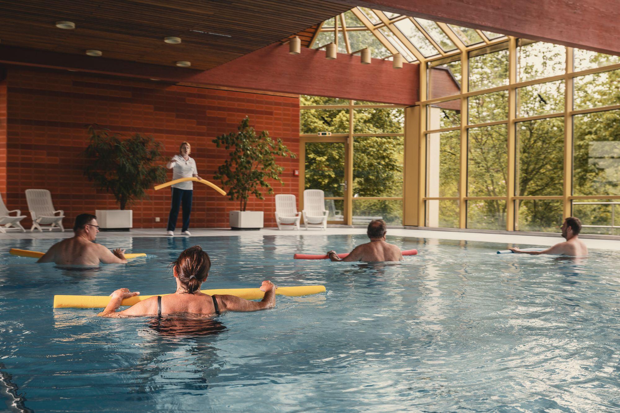 Im Schwimmbad machen mehrere Patienten Übungen mit einer Schwimmnudel unter Anleitung des Therpeuthe, der am Beckenrand steht. 