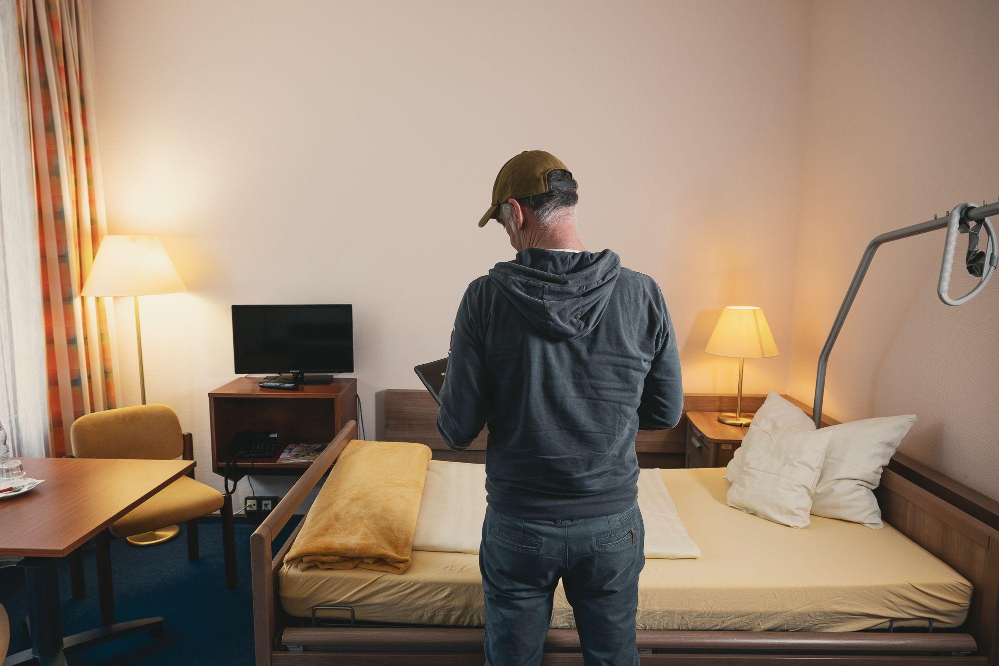 Ein Patient von hinten, der in seinem Zimmer in der Rehaklinik Lautergrund vor seinem Bett steht und in der Patientenmappe blättert.. 