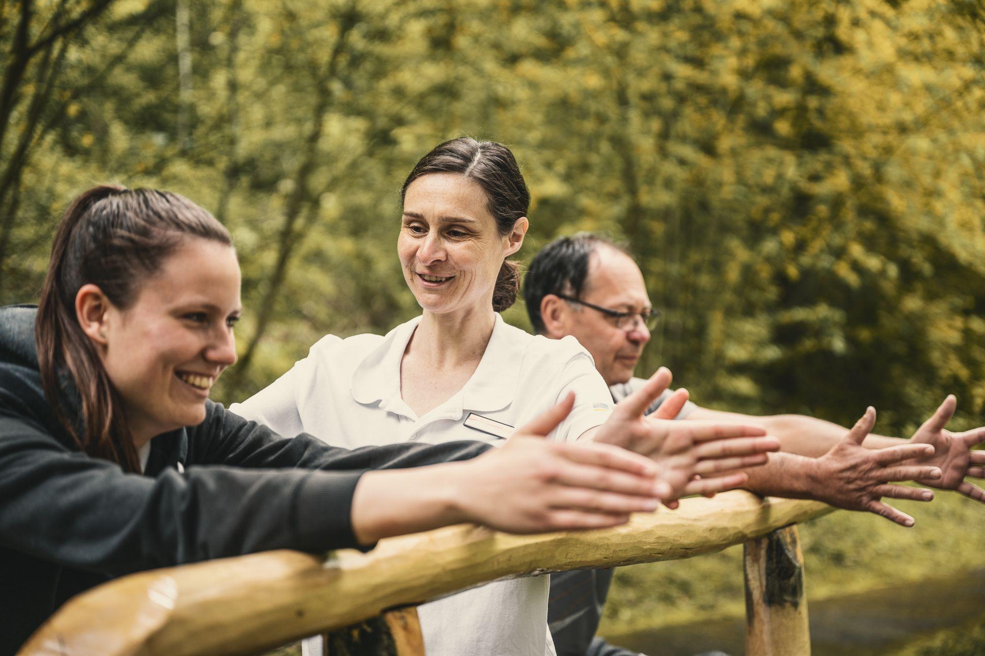 Eine Therapeutin zeigt einer Patientin eine Übung des Trimmdich-Pfades und mehrere andere Patienten schauen zu.