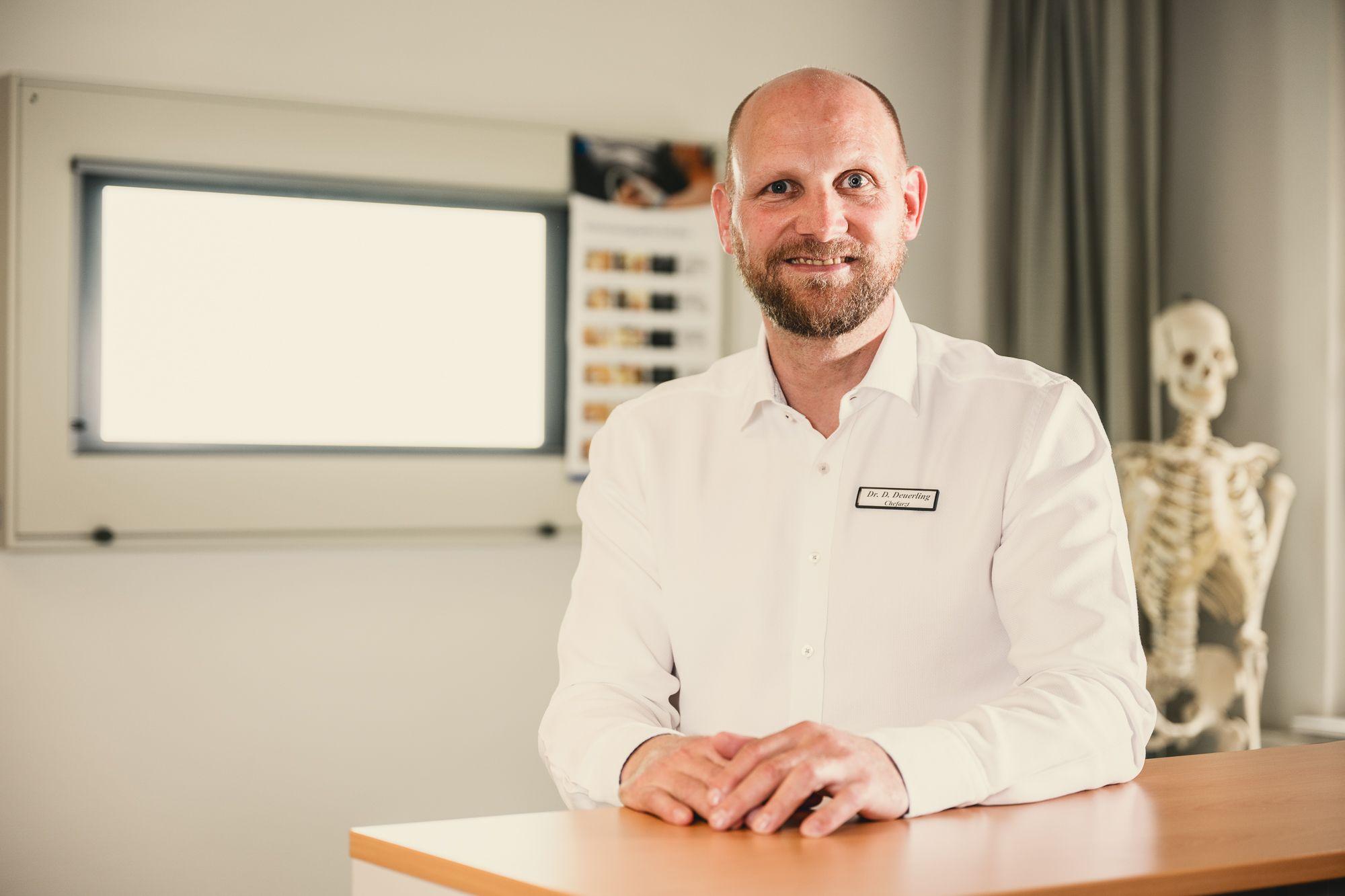 Der Chefarzt der Rehaklinik Lautergrund sitzt an seinem Schreibtisch. Im Hintergrund hängt ein Röntgenbetrachtungsbildschirm an der Wand. Hinter dem Arzt steht ein Skelett. 