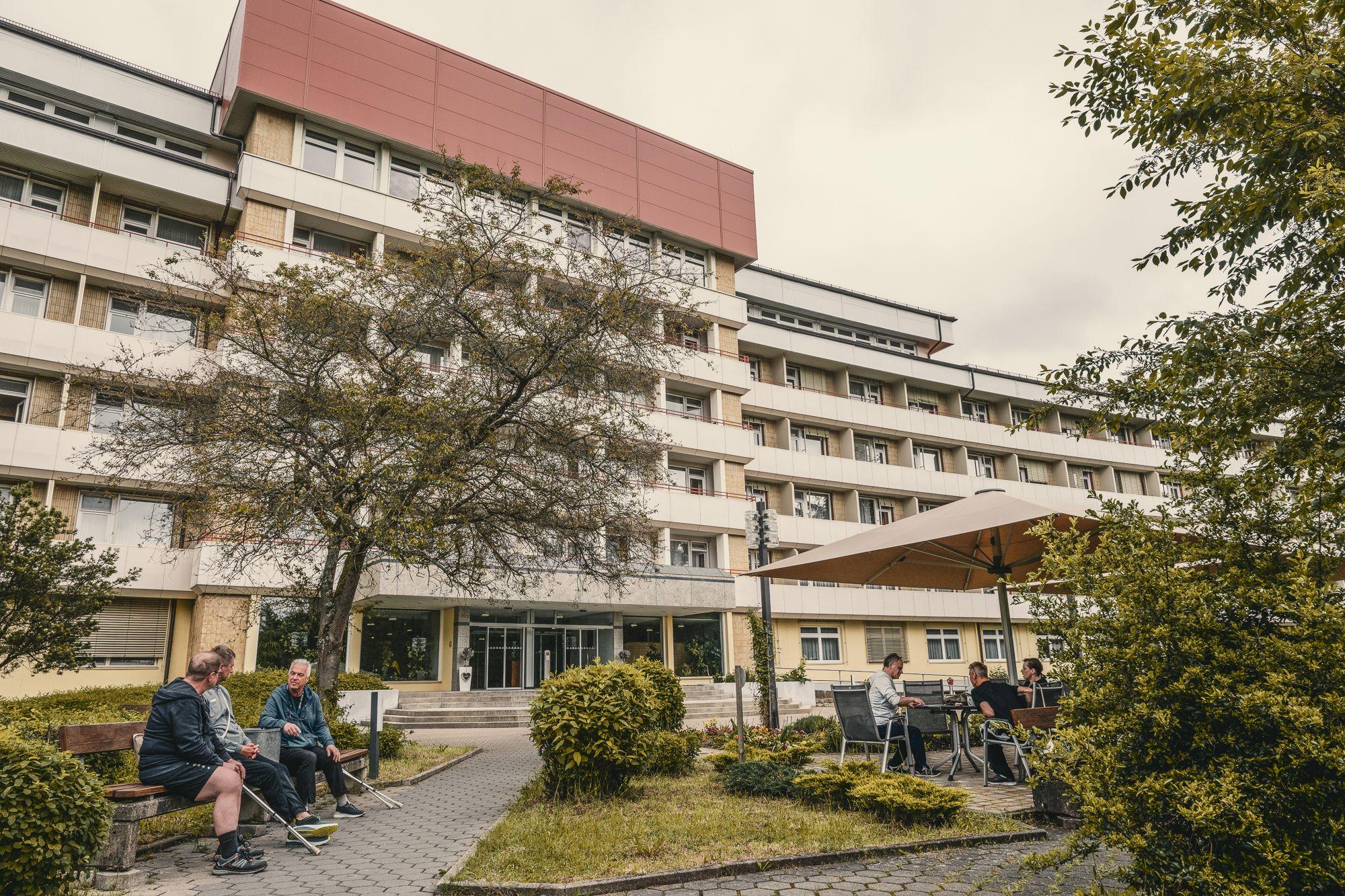 Außenansicht der Rehaklinik Lautergrund. Im Vordergrund sitzen mehrere Menschen auf Bänken und unter einem Sonnenschirm. 