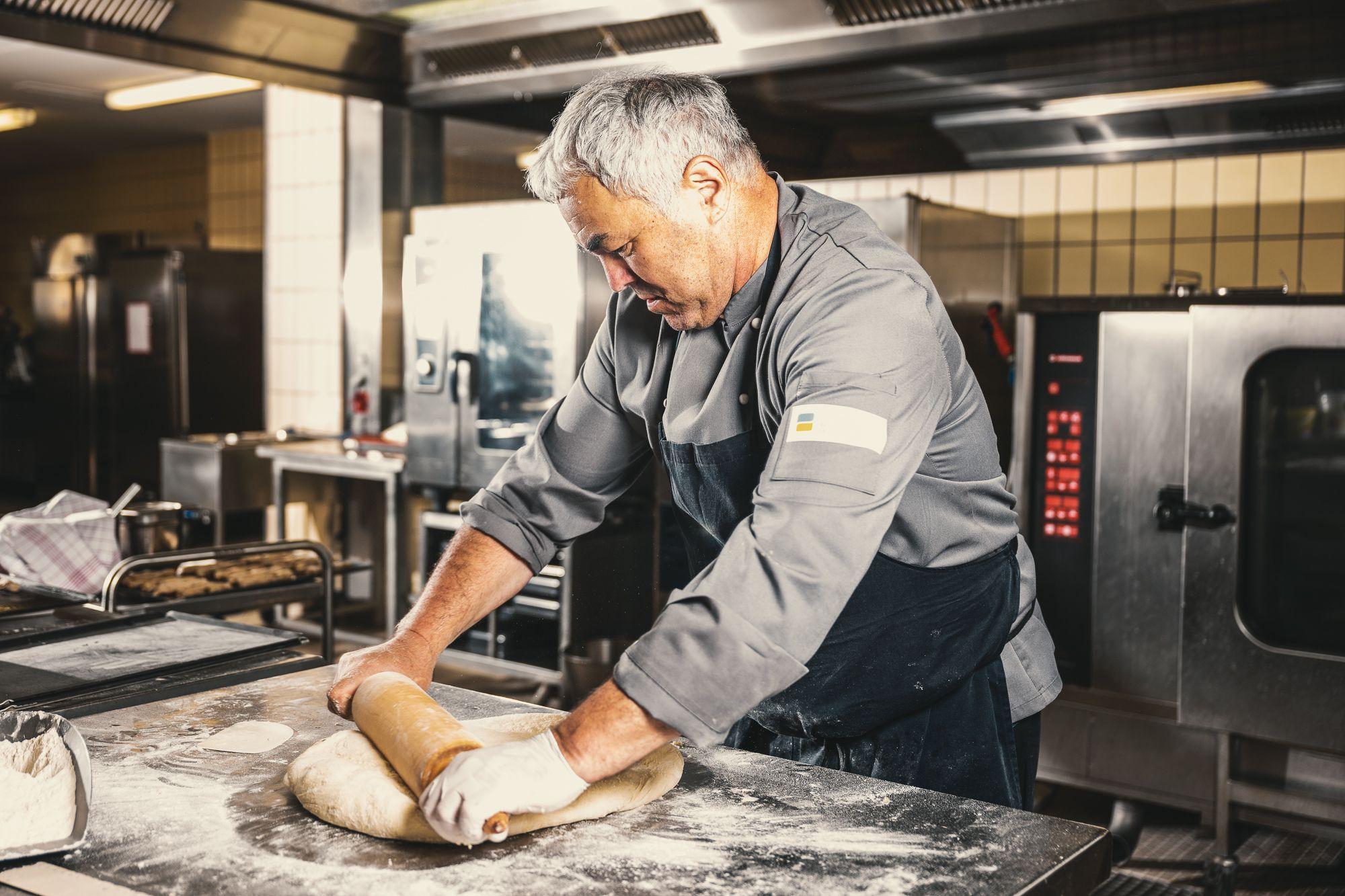 Ein Bäcker in grauem Hemd und schwarzer Schürze rollt Brotteig mit einem Nudelholz. 